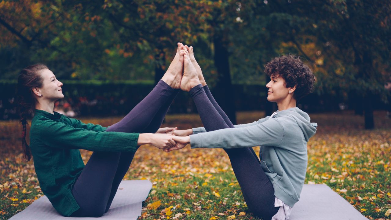 heros-img Two women practicing partner yoga outdoors on a fall day, surrounded by colorful leaves.